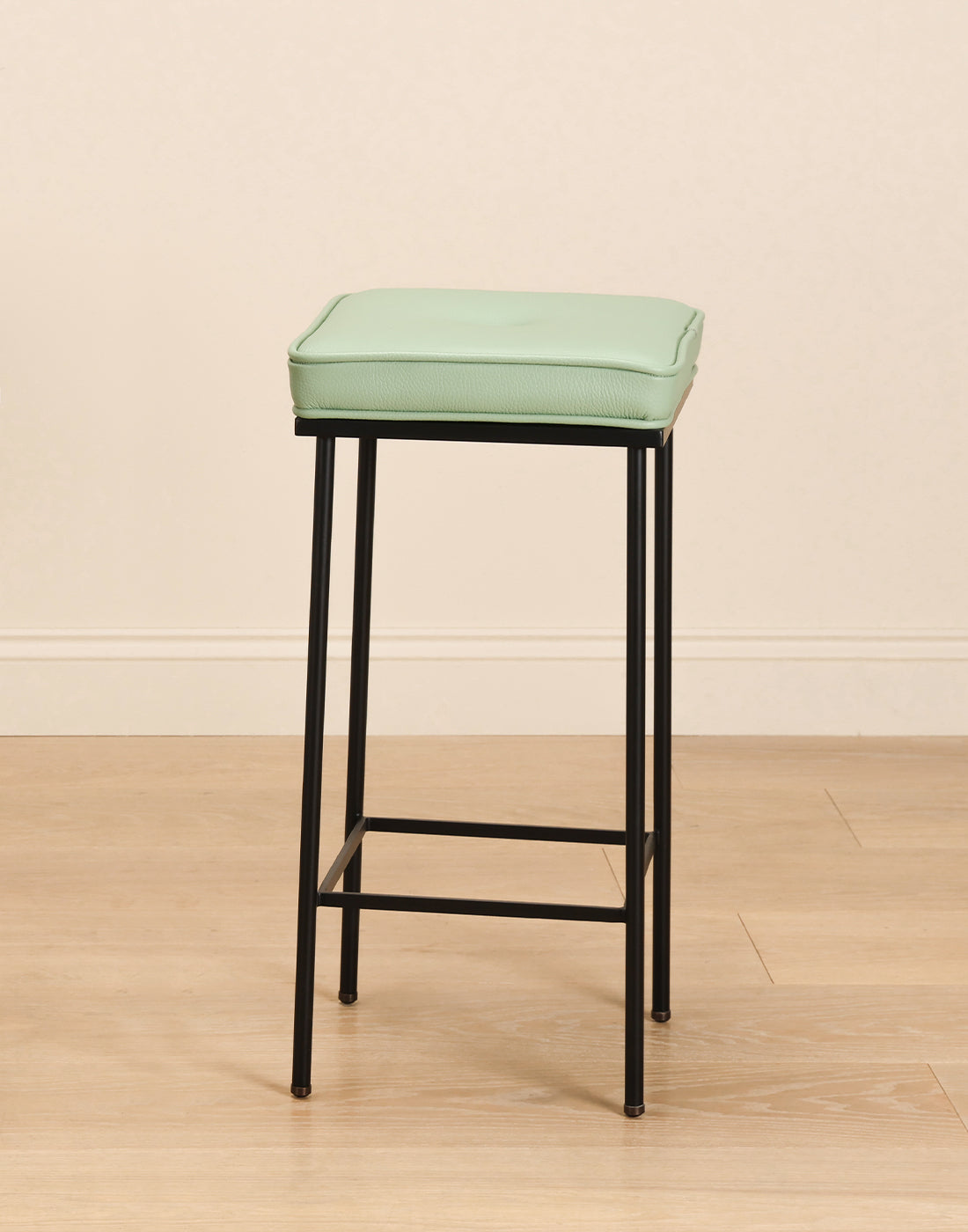 Bar stool with a green cushion and black steel frame on a wooden floor and beige wall background.