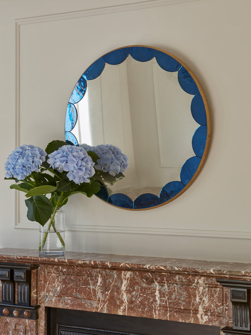 Round mirror with blue decorative frame on a marble fireplace mantel with flowers.