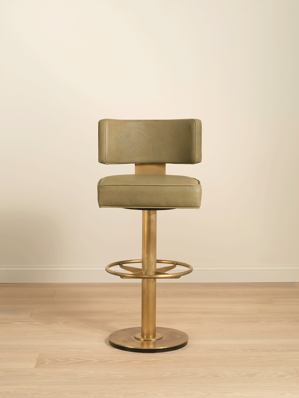 Green bar stool with brass base on a wooden floor against a beige wall.