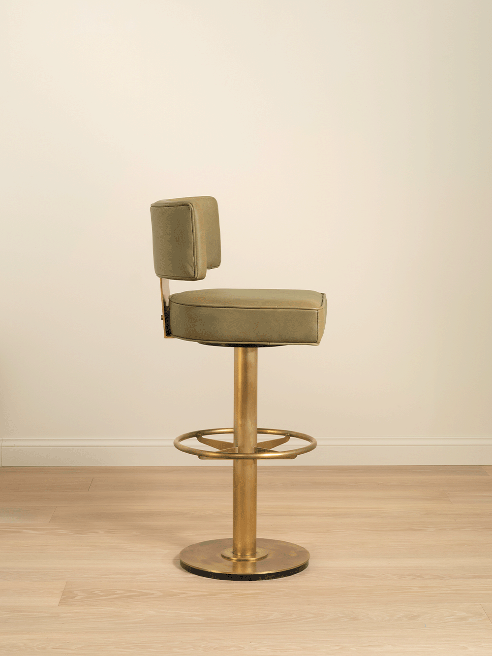 Green bar stool with brass metal base on a wooden floor against a beige wall.