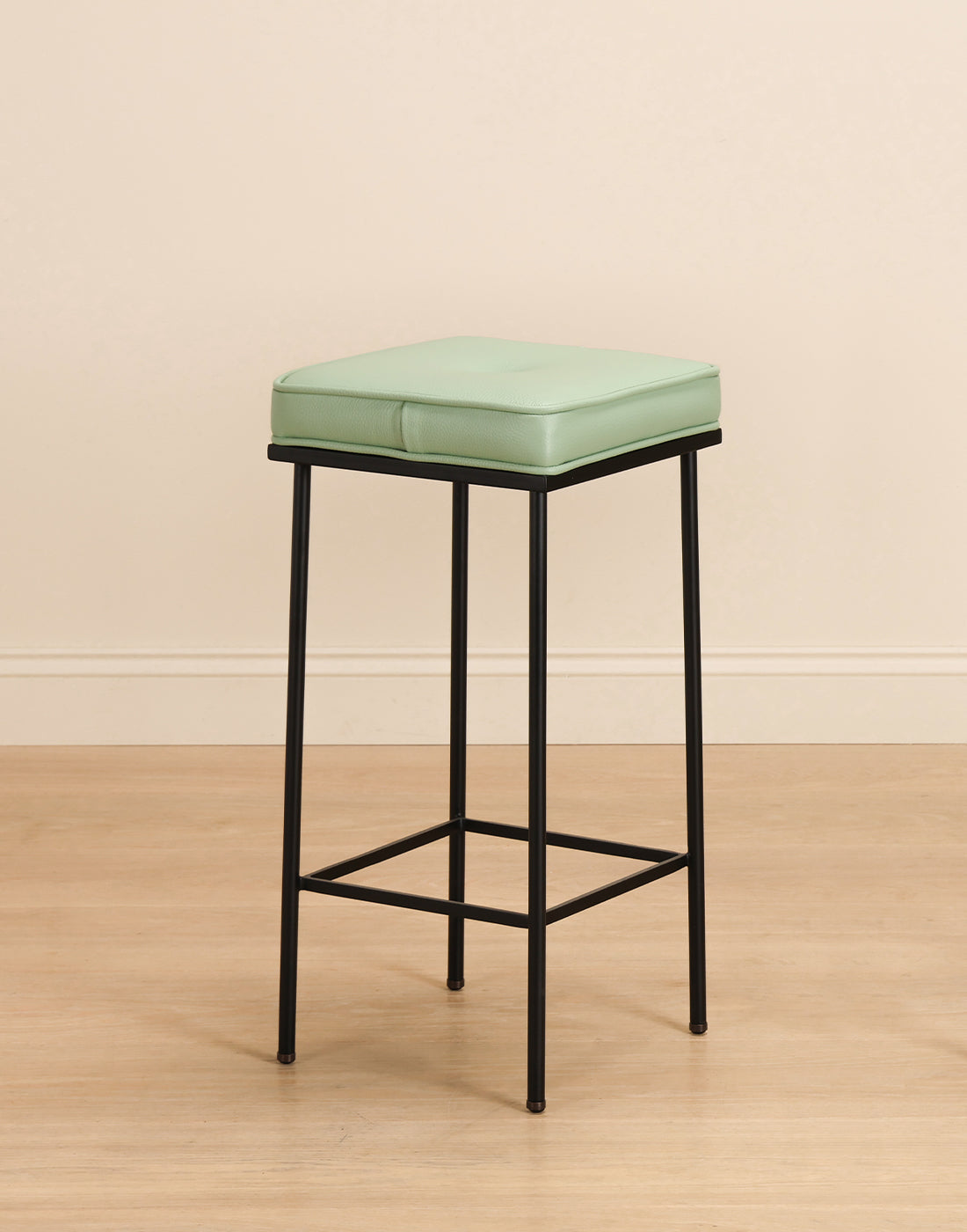 Bar stool with a green leather cushion on a wooden floor and against a beige wall.  background