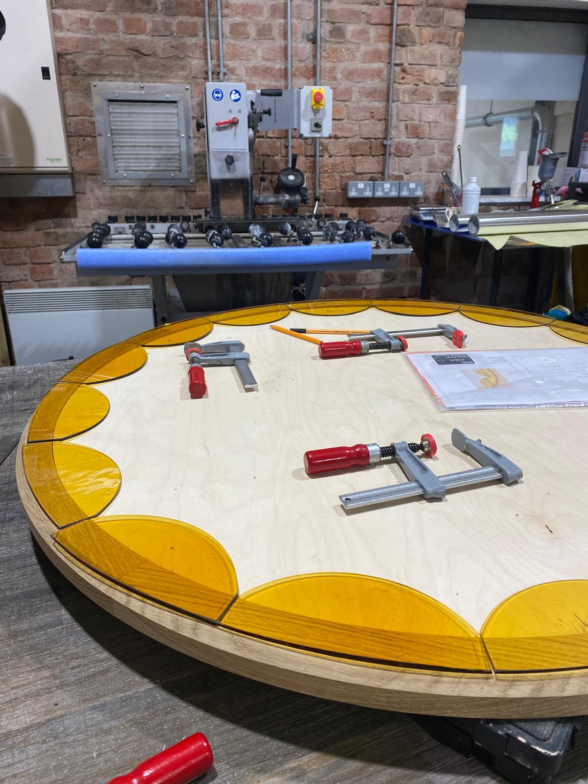 Round wooden mirror frame with yellow glass border and clamps on a workshop table.