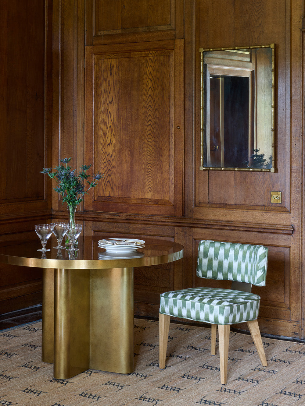Circular dining table with a green and white patterned dining chair against wooden walls with a mirror.