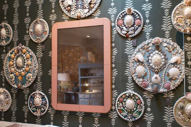 A rectangular wall mirror with a dusty pink frame, mounted on a dark green wallpapered wall decorated with numerous intricate seashell and bead medallions.
