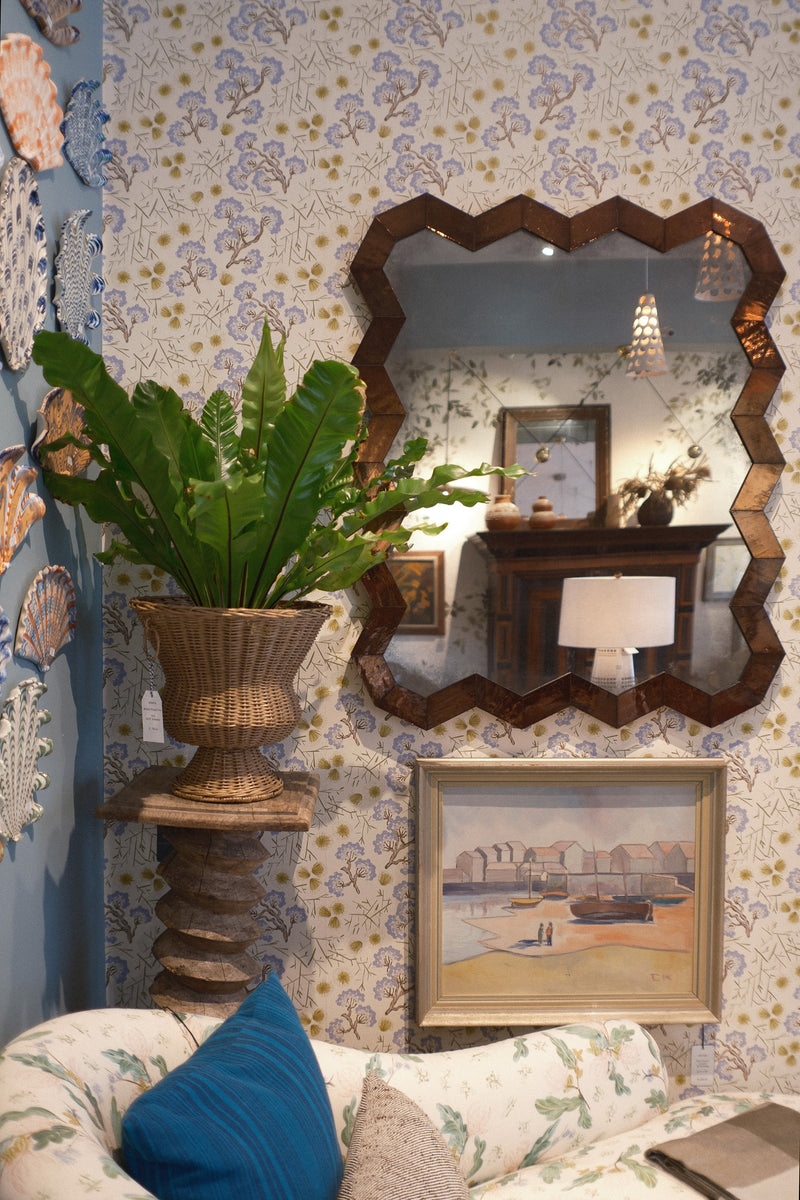 A styled interior corner featuring a large scalloped dark wood mirror on a patterned wallpaper wall, a potted green fern in a woven urn-style planter on a turned wood pedestal and a framed landscape painting hanging above a floral sofa with blue walls.