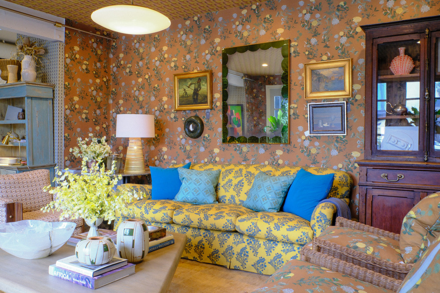 A cozy living room featuring a yellow and blue floral patterned sofa with bright blue pillows, set against orange-brown patterned wallpaper. The room is furnished with a wooden hutch, a patterned armchair and a coffee table with decorative items.