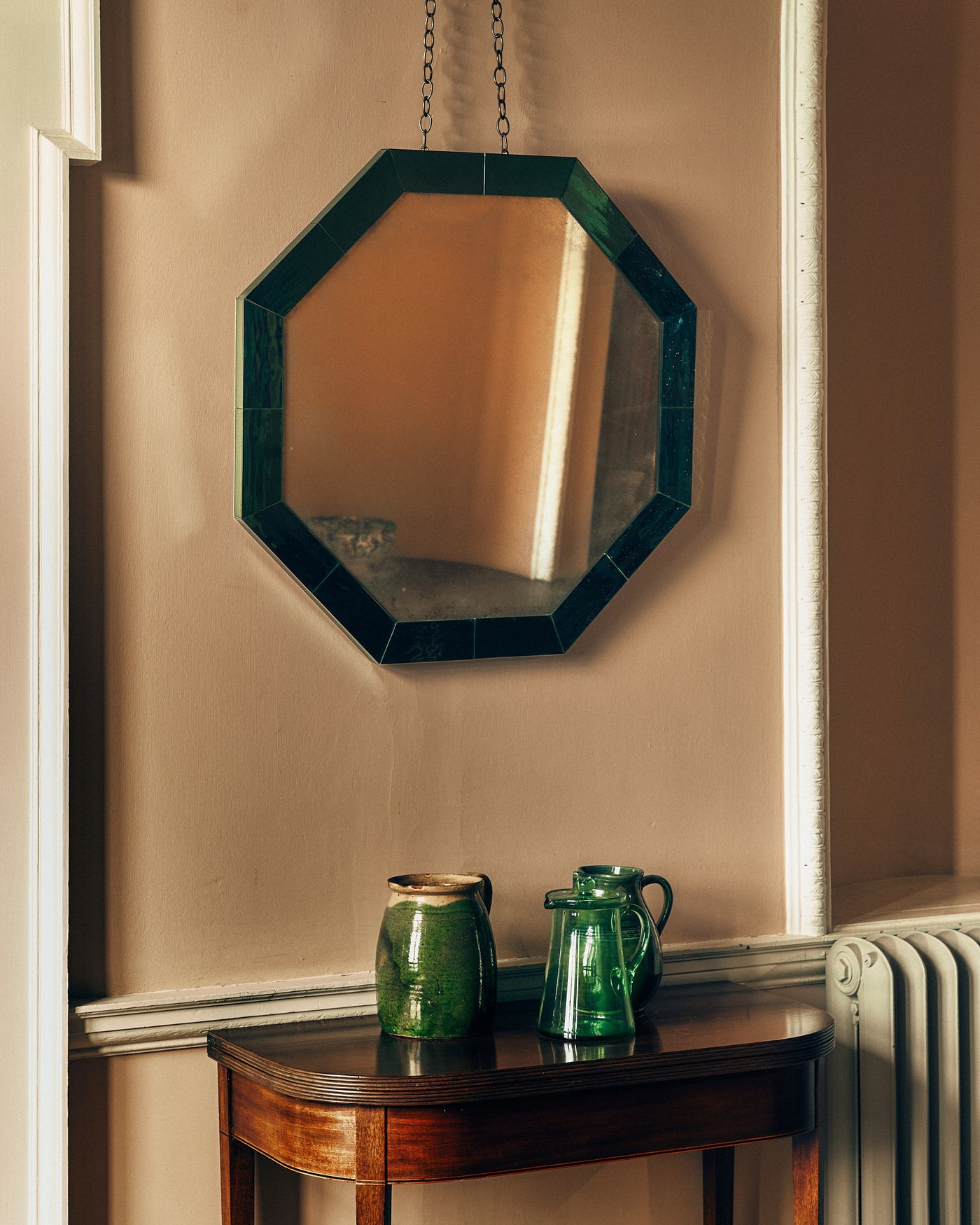 Octagonal wall mirror with a green frame with a wooden console and vases below.