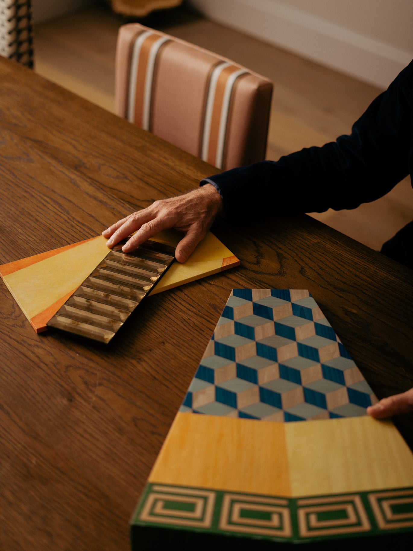 A person's hands are shown holding and displaying a set of decorative wooden sample boards with various patterns and colours, including geometric designs and wood grain textures, on a dark wooden table.