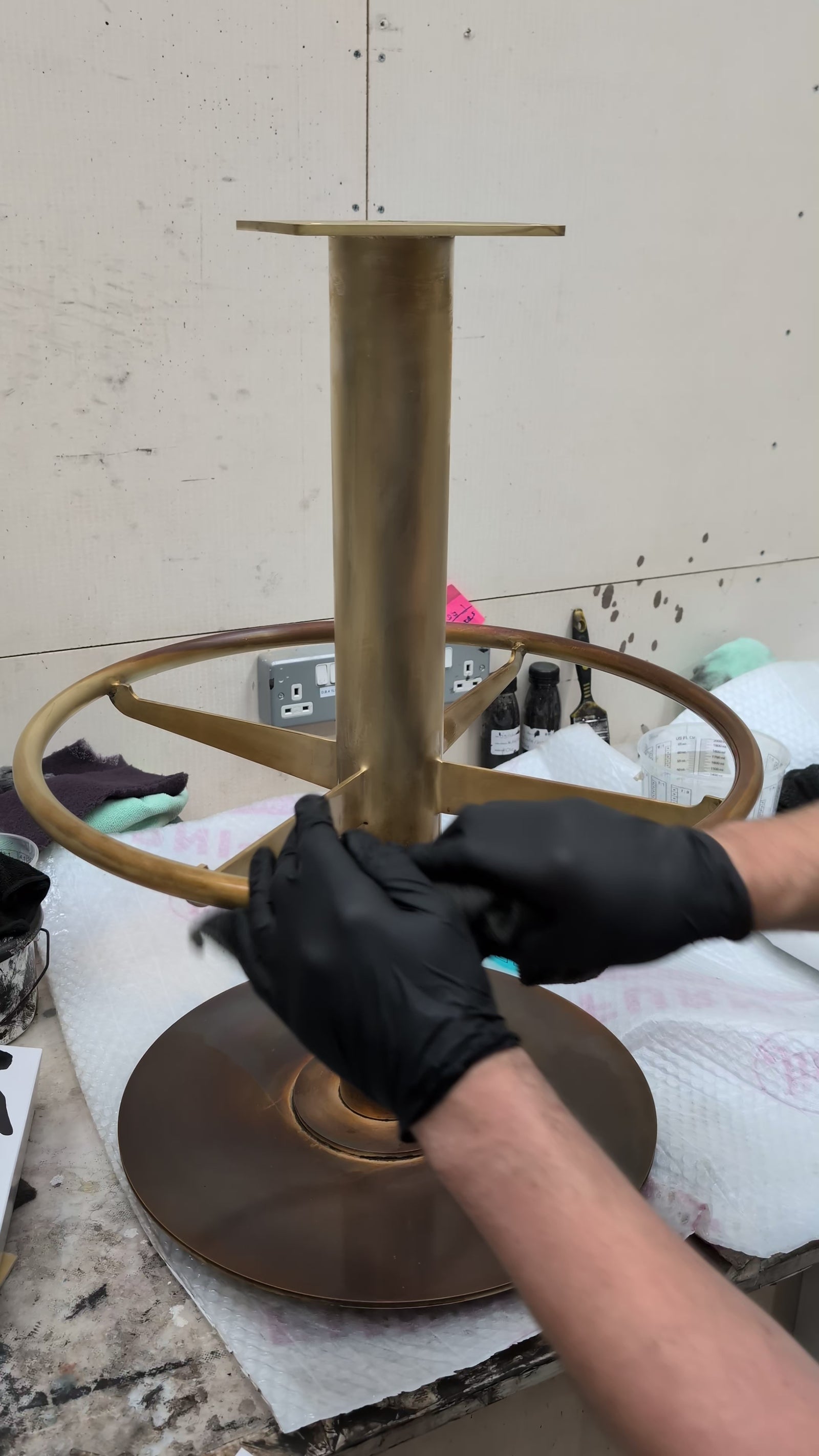Person applying a finish to a brass bar stool base in a workshop setting.